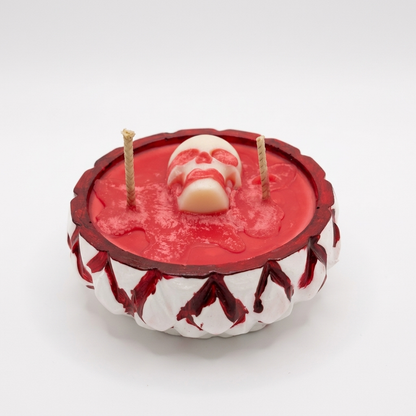 A Halloween-themed candle with a red wax base, a skull-shaped wick holder in the center, and two upright wicks, set in a white and red decorative base against a plain white background.